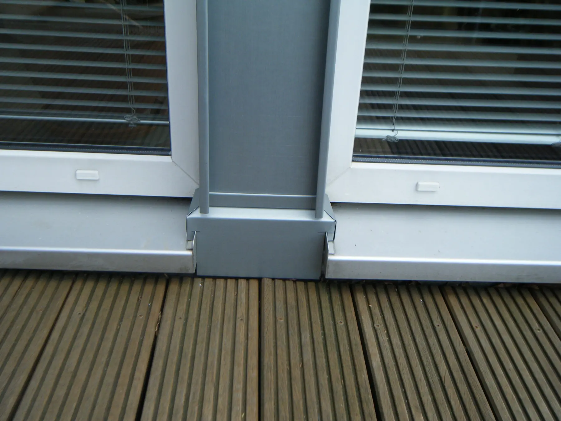 Grau beschichtete Blechverkleidung an einem Pfeiler zwischen zwei Fenstertüren, davor Holzdielen.