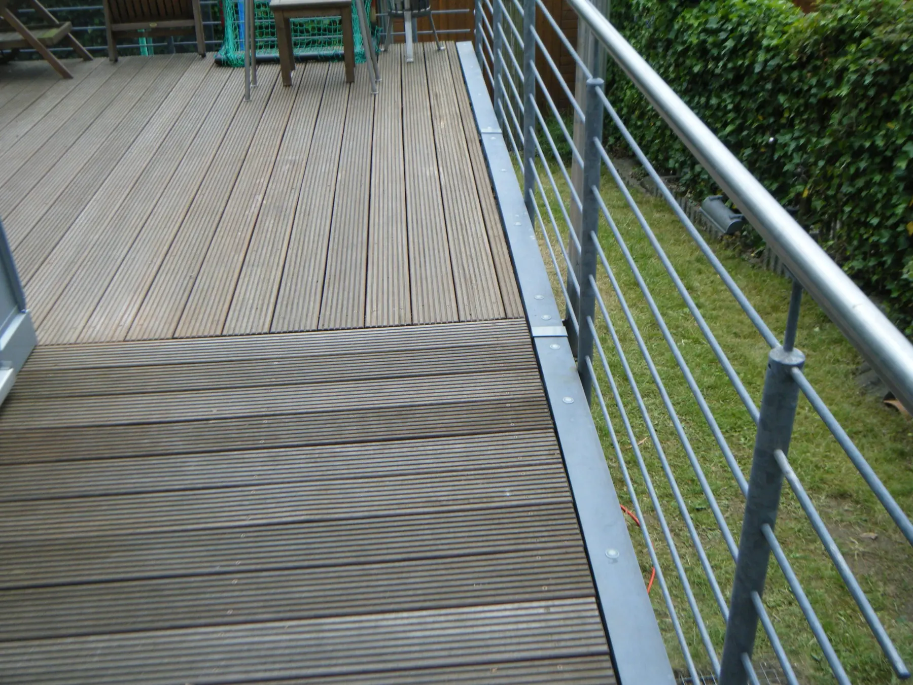 Terrasse mit Holzdielen, Randabschluss aus Metall und Stabgeländer über einem Garten.