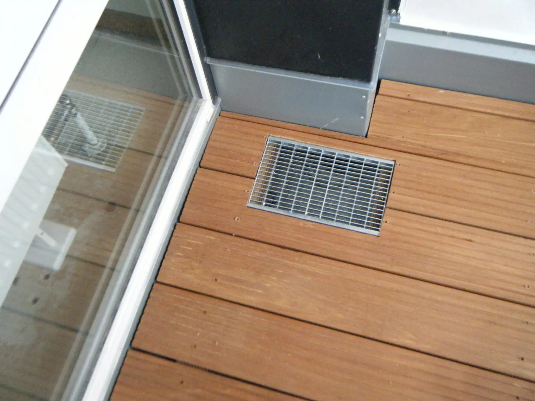 Terrassendielen aus Holz mit eingelassenem Ablaufrost vor einer Glastür, daneben eine Pfeilerverkleidung aus Blech.