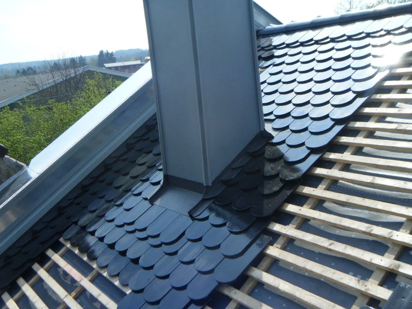 Neue Eindeckung mit dunklen Biberschwanzziegeln rund um einen metallverkleideten Kamin, daneben freie Lattung.