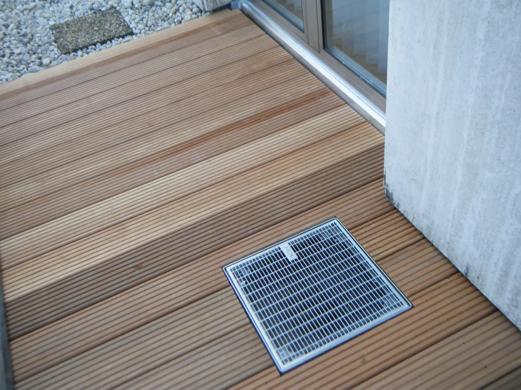 Holzterrasse mit Stufe und quadratischem Ablaufrost vor einer Fenstertür, links ein Kiesbett.