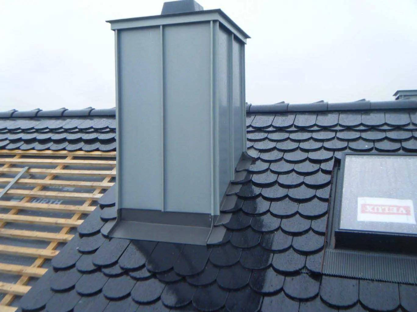 Steildach mit dunklen Biberschwanzziegeln, metallverkleidetem Kamin und Dachfenster.