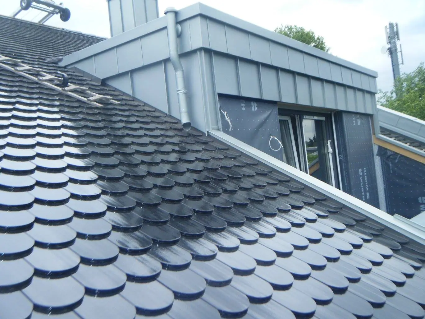 Gaube mit grauer Metallverkleidung und Fallrohr auf einem Dach mit dunklen Biberschwanzziegeln.